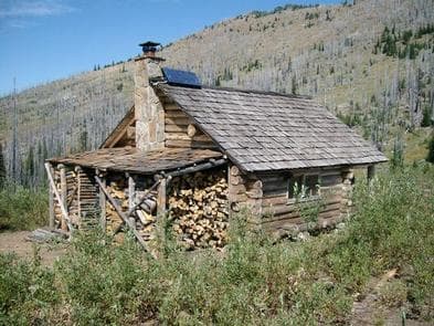 Log cabin set in brush, a full woodshed overlooking a hillside blanketed with silvered conifer snags.