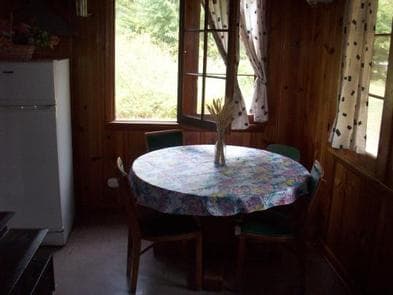 Round table covered with floral cloth, set with dried flower arrangement, circled by four chairs looking through open window and white patterned curtains to sunny, forested yard.