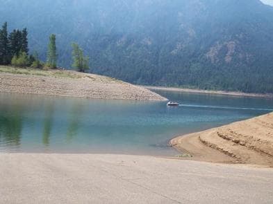 View of boat ramp leading into lake with sloped earthen banks, looking beyond to a small boat and tree covered slopes above high water line.