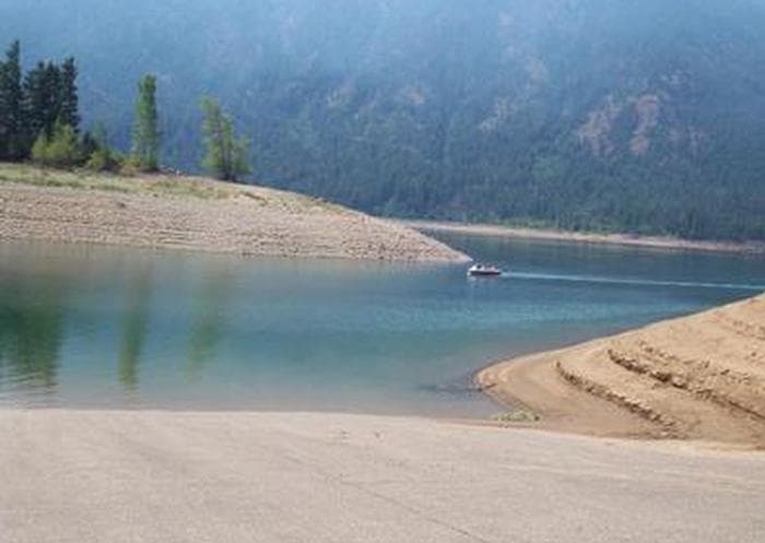 View of boat ramp leading into lake with sloped earthen banks, looking beyond to a small boat and tree covered slopes above high water line.