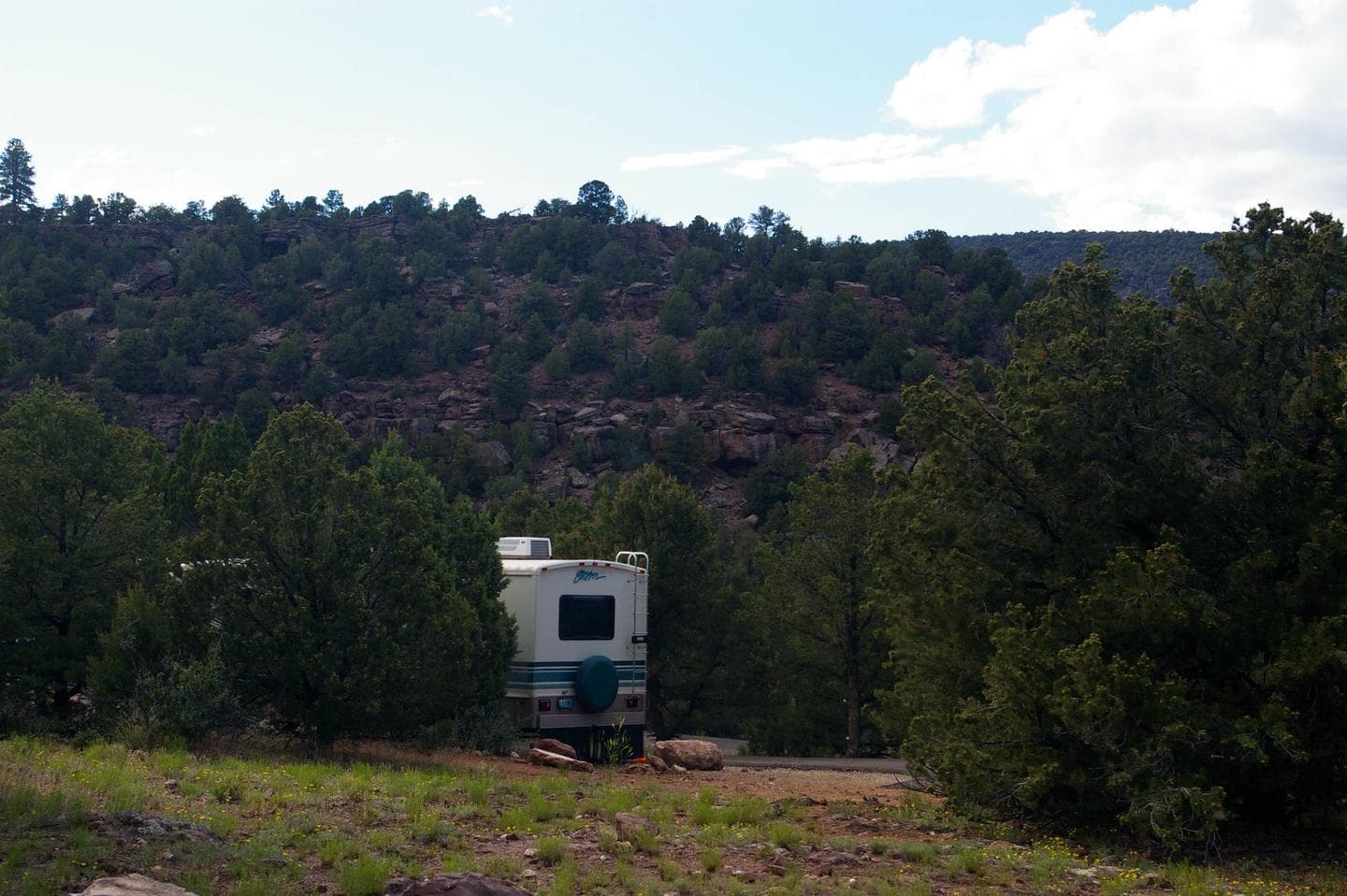 A large motor home is parked in a camp site. There are trees lined on both sides and a hillside with red rocks and trees can be seen from the site.