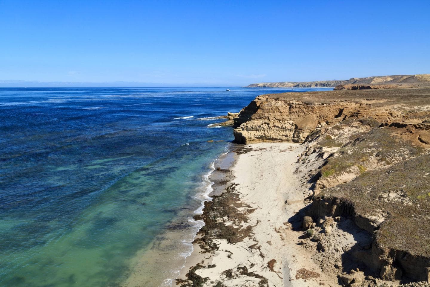 Sandy beach with ocean, small waves and steep coastal bluffs covered in dry grass.