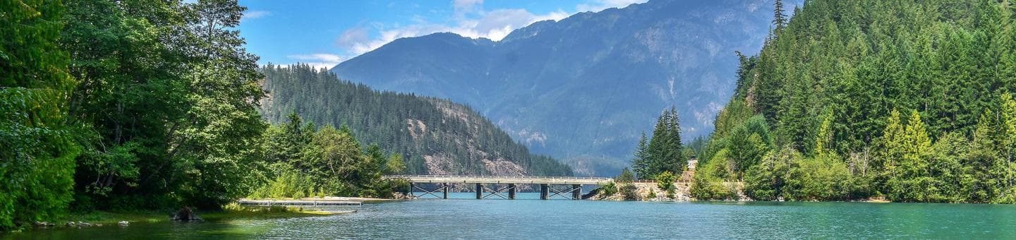 View of a lake with lush trees on either side, highway bridge and a mountain in the background with blue sky