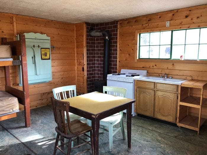 Cabin interior showing gas range, wood stove, table and dry sink.