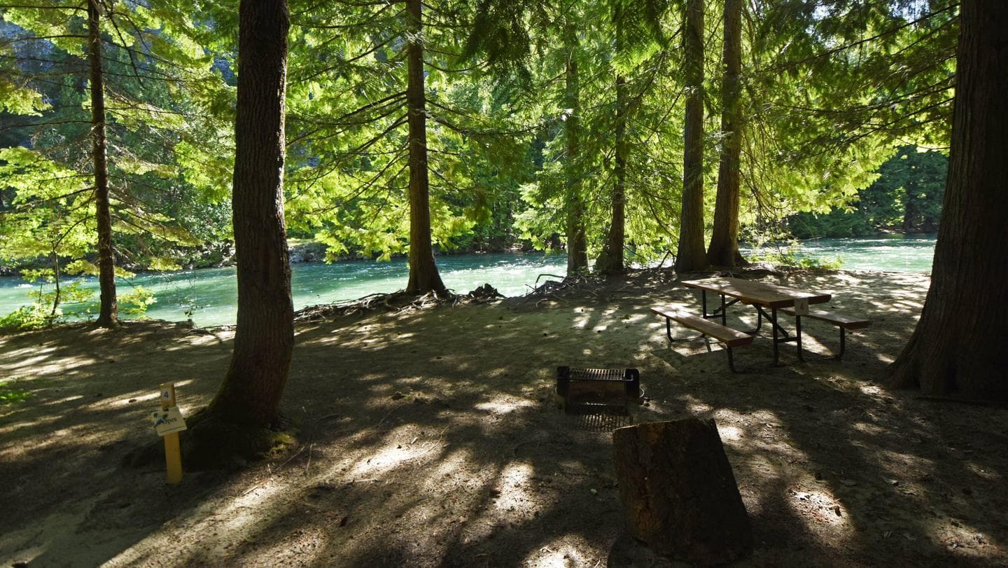 Picnic table and fire grill with trees and river in the background.