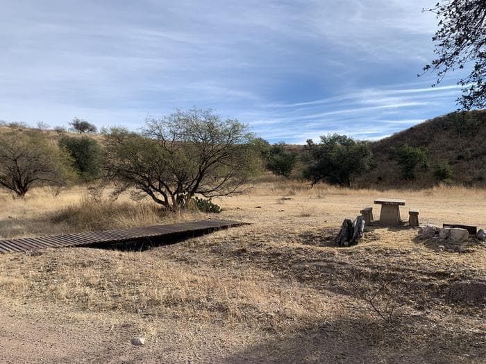 A small flat foot bridge crosses a shallow wash to a wide open space with one picnic table