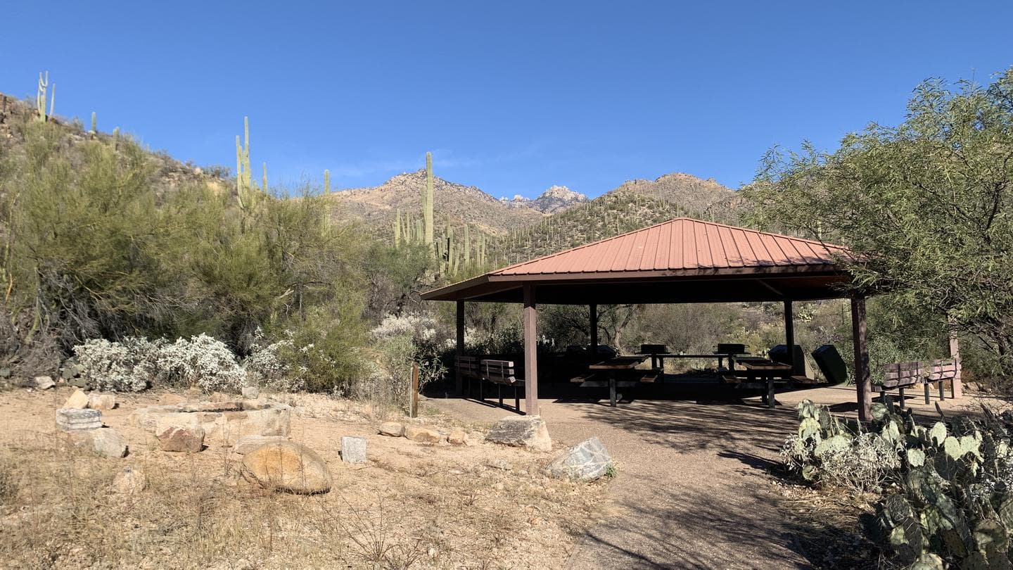 A covered ramada with picnic tables, grills, trashcans, and bear-safe contatiners. Stone fire pit immediately to the left.