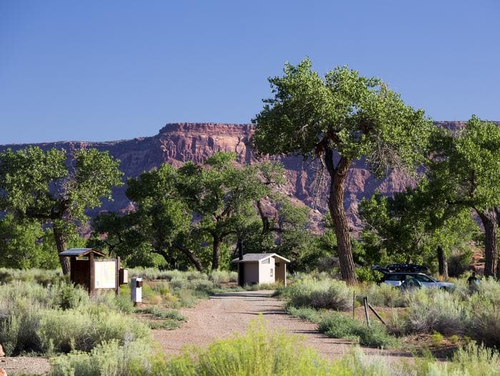 A registration kiosk and and vault toilet under Cottonwood trees with red rock cliffs in the background.