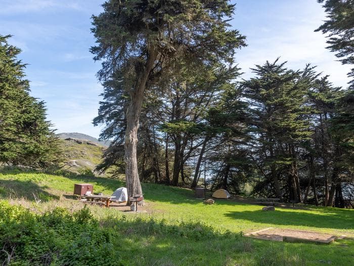 Side view of Bicentennial Campground. In the foreground is Site 1, to the left is Site 2, and in the background is Site 3. Through the cypress trees surrounding the campground, you can see the hills of the headlands.