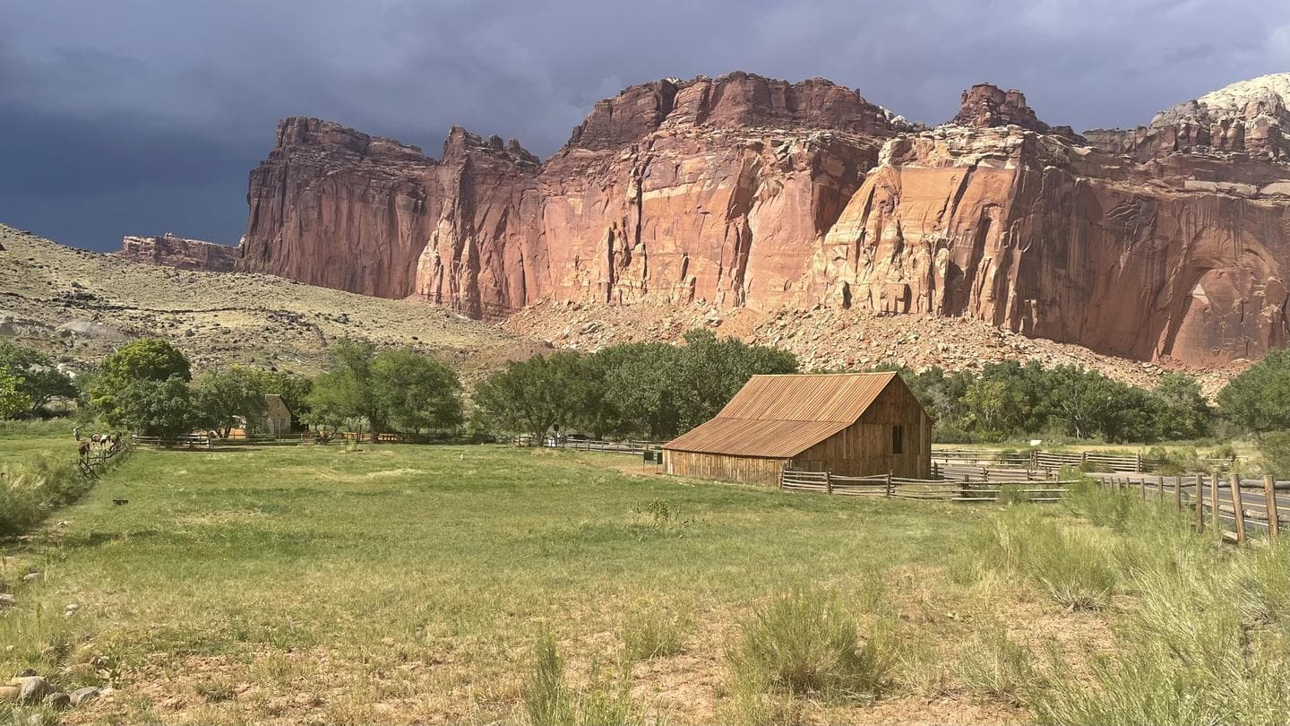 A old barn sites in a grass field. A red rock cliff is the background.