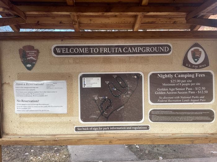 A Bulletin Board with 8 signs in it. The signs are as follows, starting at the top center and moving clockwise: "Welcome to Fruit Campground"; "U.S. Fee Area"; "Night Camping Fees. $25.00 per site; Maximum of 8 people per site. Golden Age/Senior Pass- $12.50; Golden Access/Access Pass- $12.50. No discount with National Parks and Federal Recreation Lands Annual Pass"; Privacy Act sign; Map of the campground; "See back of sign for park information and regulations"; "Have a Reservation? Refer to the campground map and proceed to your campsite. Don't know your campsite number? Go to the visitor center to connect to free public Wi-Fi to access your reservation confirmation. Wi-Fi is available 24/7. No Reservation? All sites must be reserved through Recreation.gov. Go to the visitor center to connect to free public Wi-Fi to reserve a site. Wi-Fi is available 24/7. Visitor center staff cannot reserve the site for you. The top right corner of the same sign reads as follows: "Seasons. Loops A & B: Open year-round. Loop C: Open March 1st- End of November."; The NPS Arrowhead.