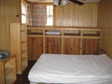 Horsethief Cabin bedroom with full bed, bookcases at the foot, two windows, ceiling fan and cabinets on back wall.